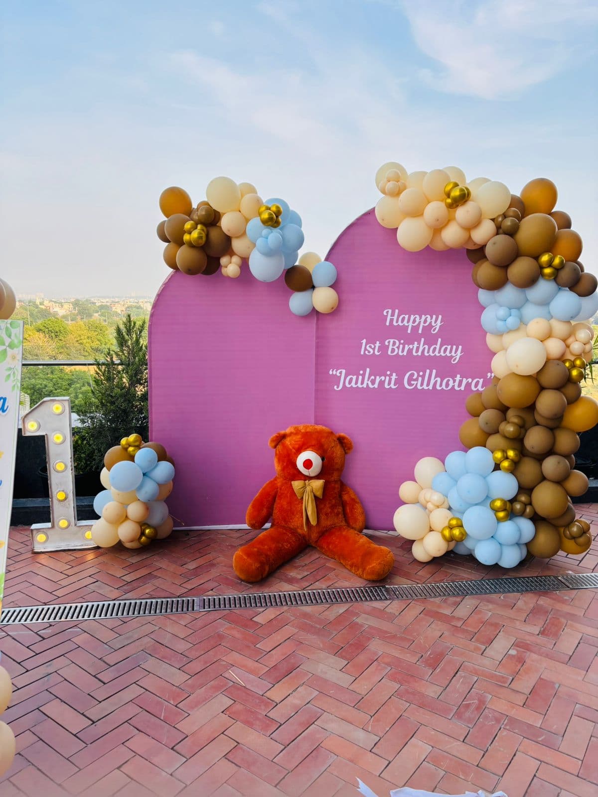 Pastel first-birthday arch with a teddy and a lit number 'One'