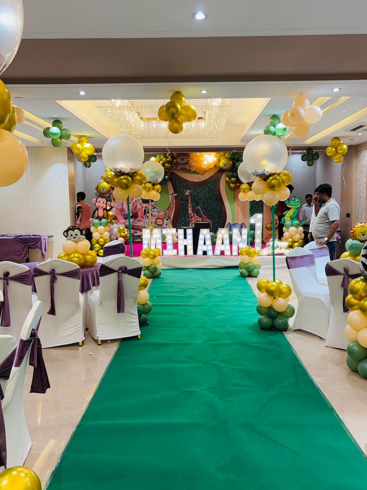 Jungle-themed first-birthday setup with a green walkway and animal cut-outs in a banquet hall